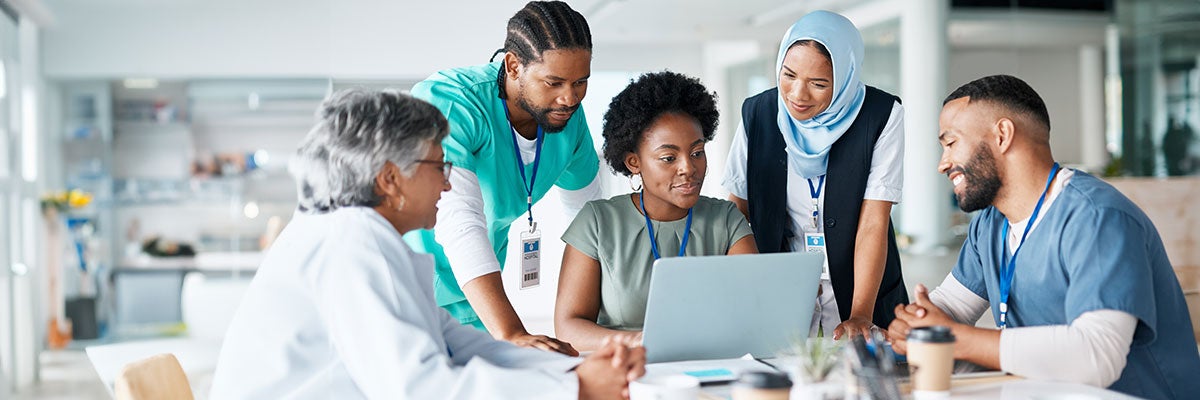 A diverse team of men and women sit and stand gathered around a laptop looking at screen